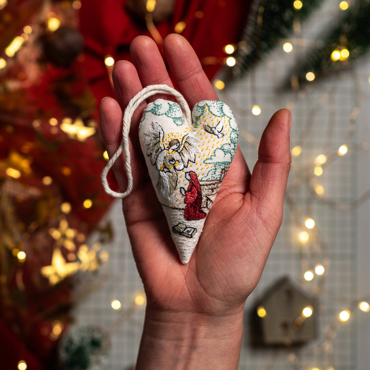Small handmade Annunciation heart ornament with white hanging cord resting in an open palm against festive Christmas lights