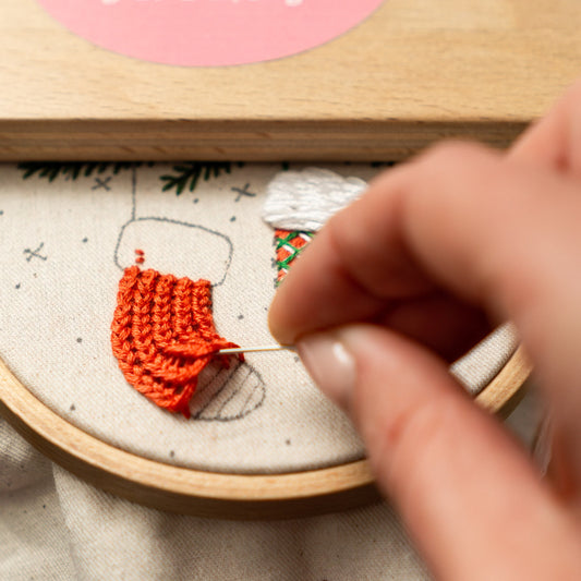 May include: Close-up of a hand embroidering a red knitted stocking on a white fabric with a wooden embroidery hoop. The stocking is partially embroidered with a green and white pattern.