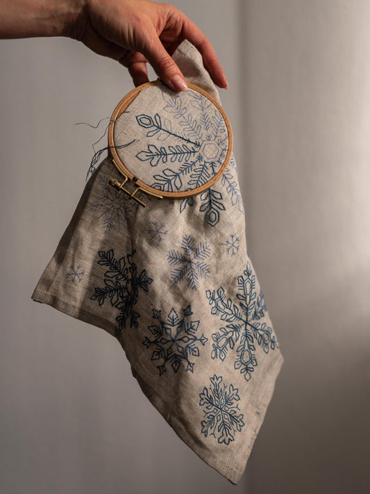 May include: A beige linen fabric with blue snowflake embroidery, held in a wooden embroidery hoop. The design features various snowflake patterns. The fabric is partially embroidered, with loose threads visible.