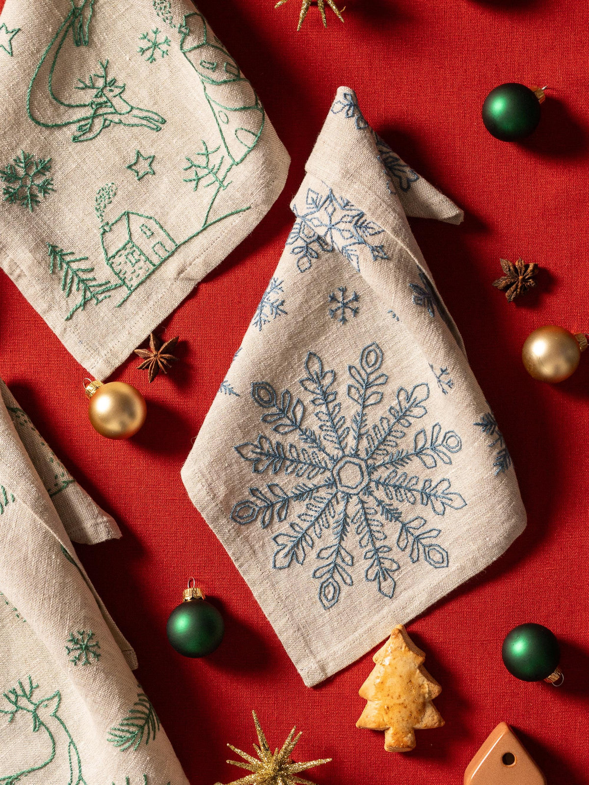 May include: Beige linen napkins with embroidered Christmas designs, including snowflakes, reindeer, and houses. The napkins are arranged on a red surface with green and gold ornaments, star anise, and a gingerbread tree.