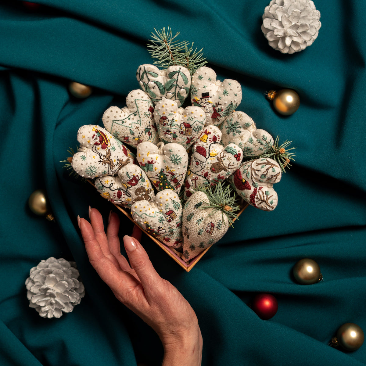 May include: A white heart-shaped ornament with a winter scene embroidered on it. The scene includes skiers, snowboarders, and trees. The ornament is hanging from a string.