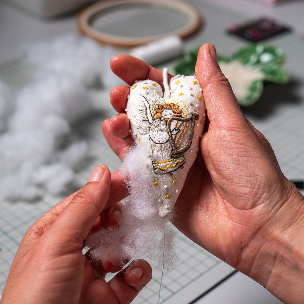 A close-up of hands stuffing a white embroidered angel-shaped ornament with polly fiber. The ornament features gold and brown embroidery of an angel with wings, set against a blurred crafting workspace background.