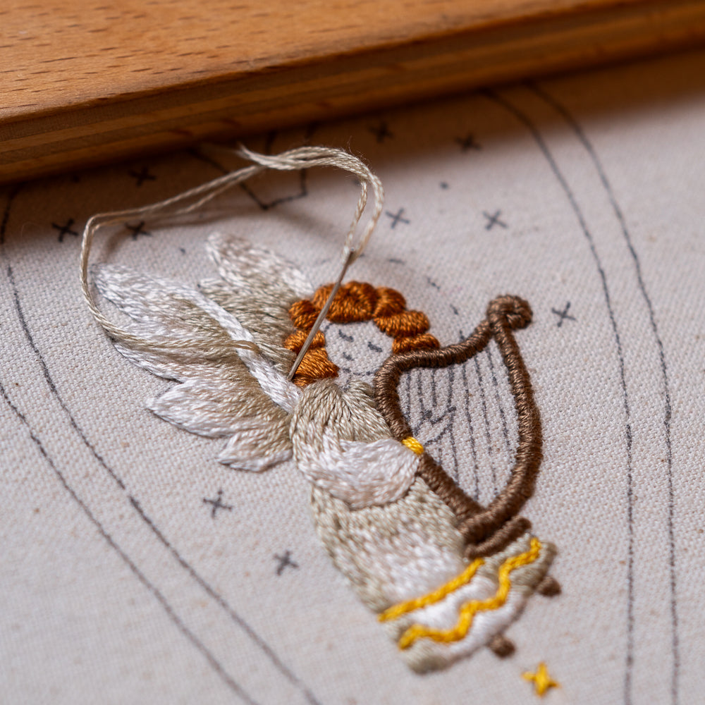 Close-up of a hand-embroidered angel figure holding a brown harp, stitched on light cotton fabric.
