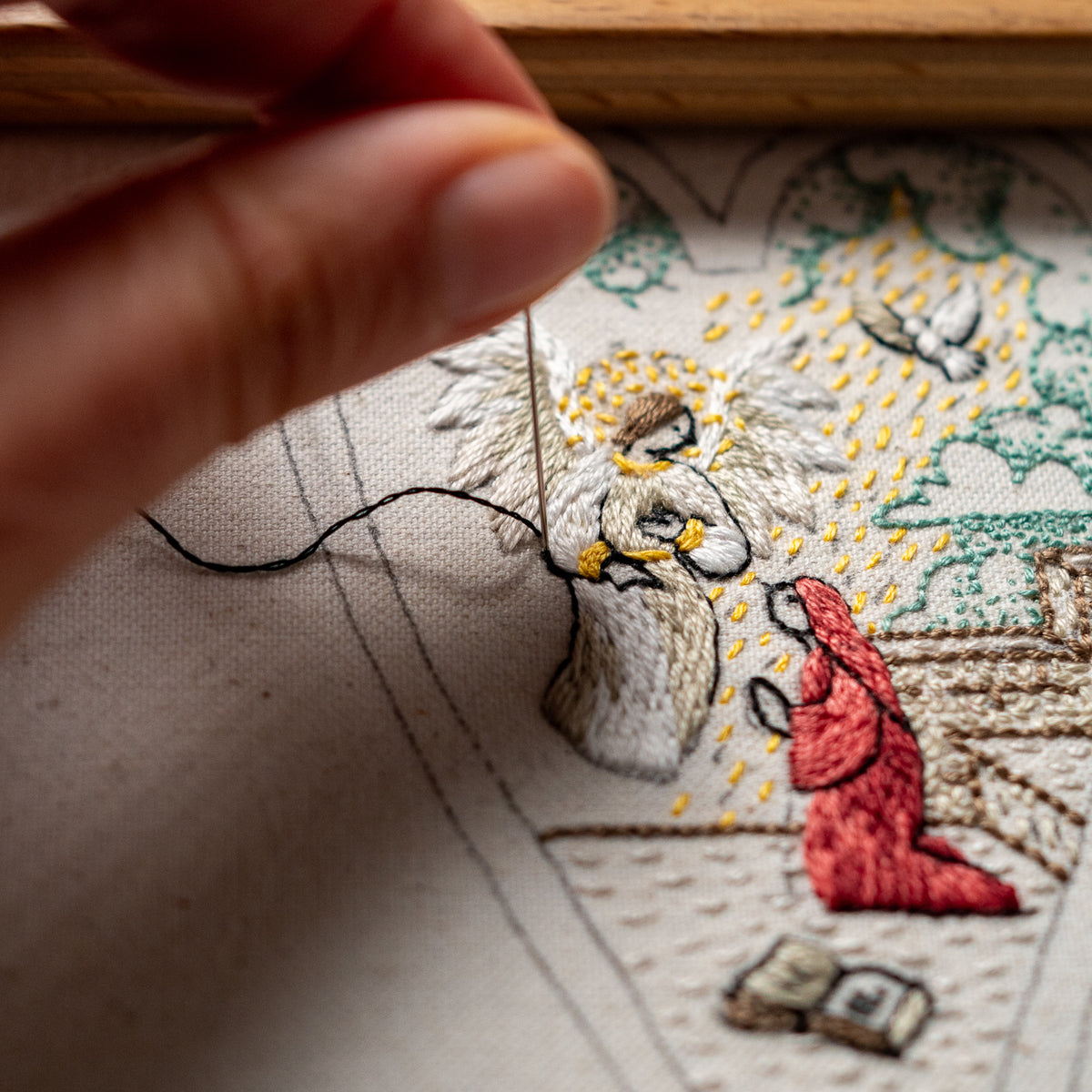 Close-up of hand embroidery in progress showing Angel Gabriel, Mary in a red robe, a dove and detailed stitching.