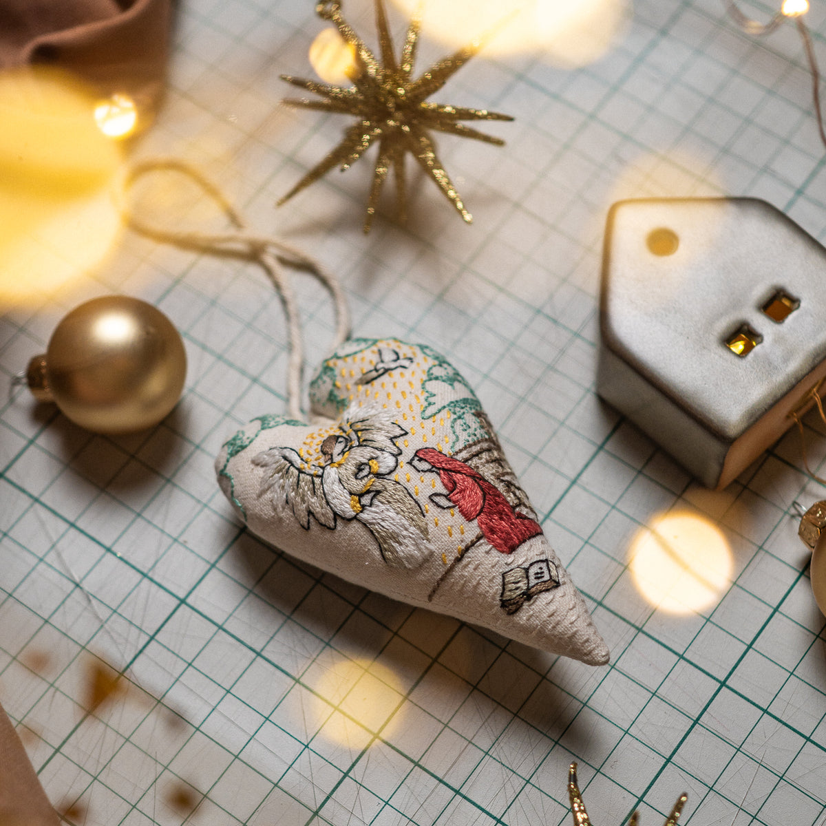 Finished Annunciation heart ornament with hanging cord, displayed with gold Christmas baubles, red fabric and fairy lights.