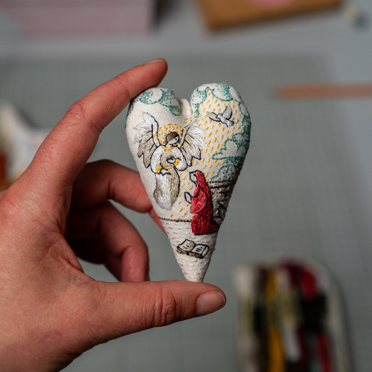 Finished handmade Nativity heart ornament held between fingers, showing Angel Gabriel, Mary and delicate embroidered details.