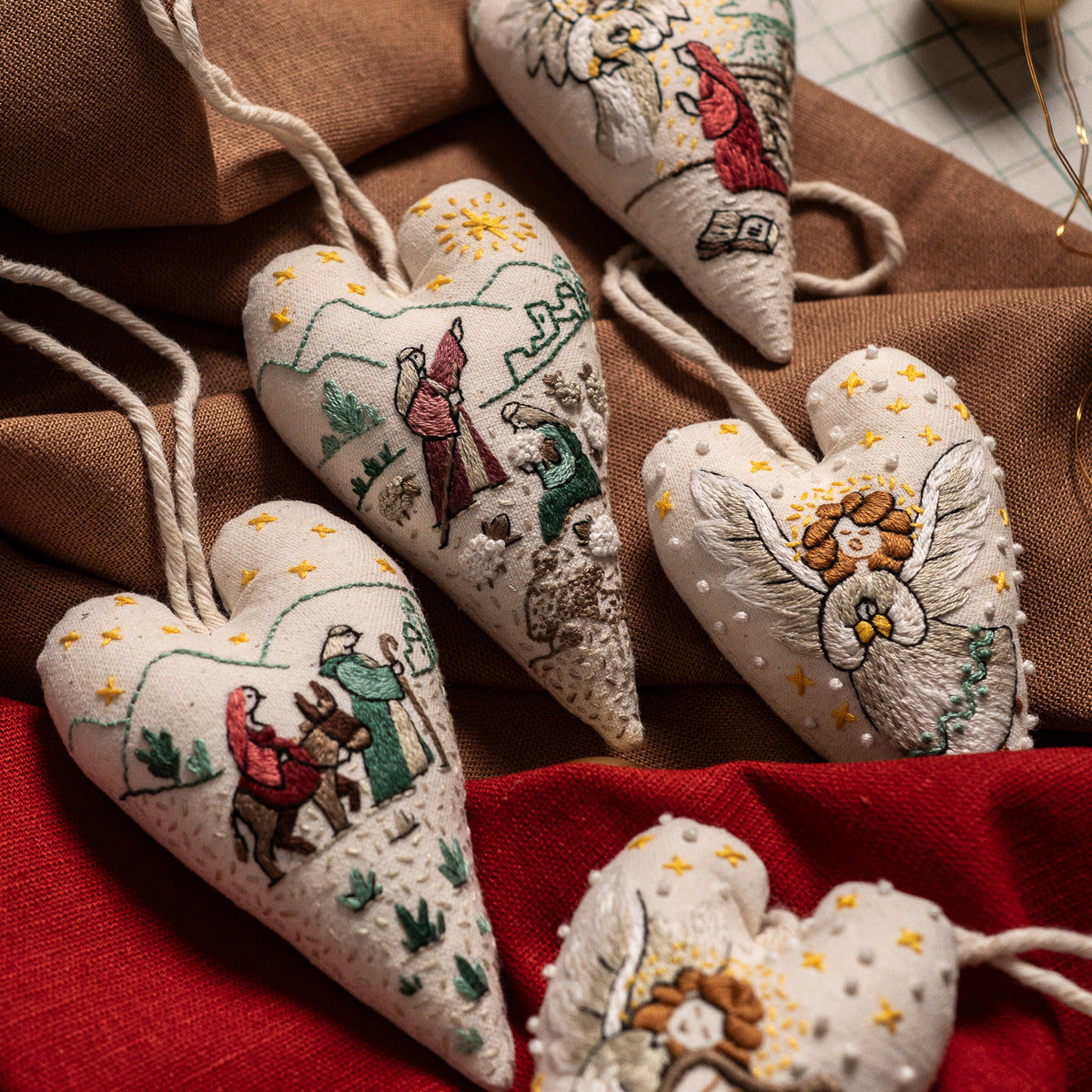 Collection of handmade Christian Christmas heart ornaments with embroidered Nativity scenes, shepherds, angels and sheep arranged on warm fabric.