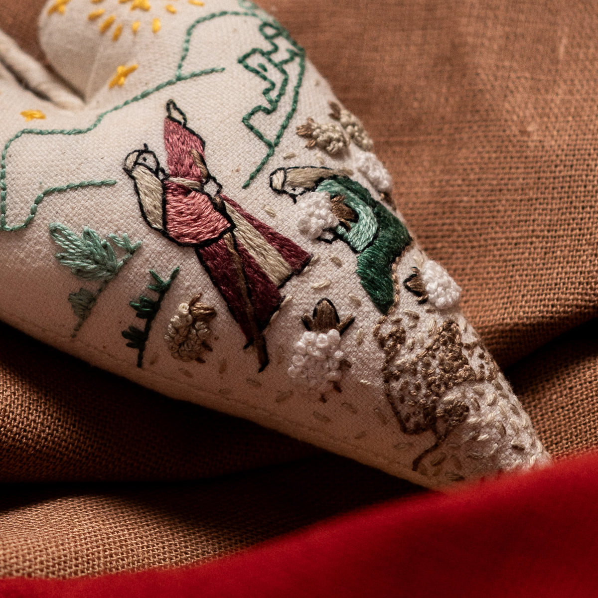 Close-up of The Shepherds of Bethlehem Christmas embroidery showing two shepherds, textured sheep, green hills and the town of Bethlehem on a handmade heart ornament.