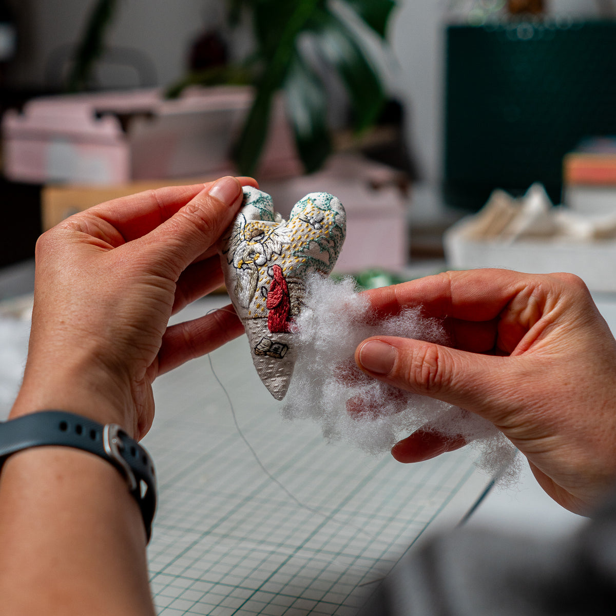 Hands adding soft stuffing to The Annunciation embroidered heart ornament during the finishing process.