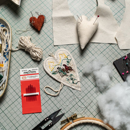 Overhead view of sewing supplies, embroidery tools, fabric hearts, and stuffing used to assemble The Road to Bethlehem Christmas ornament.