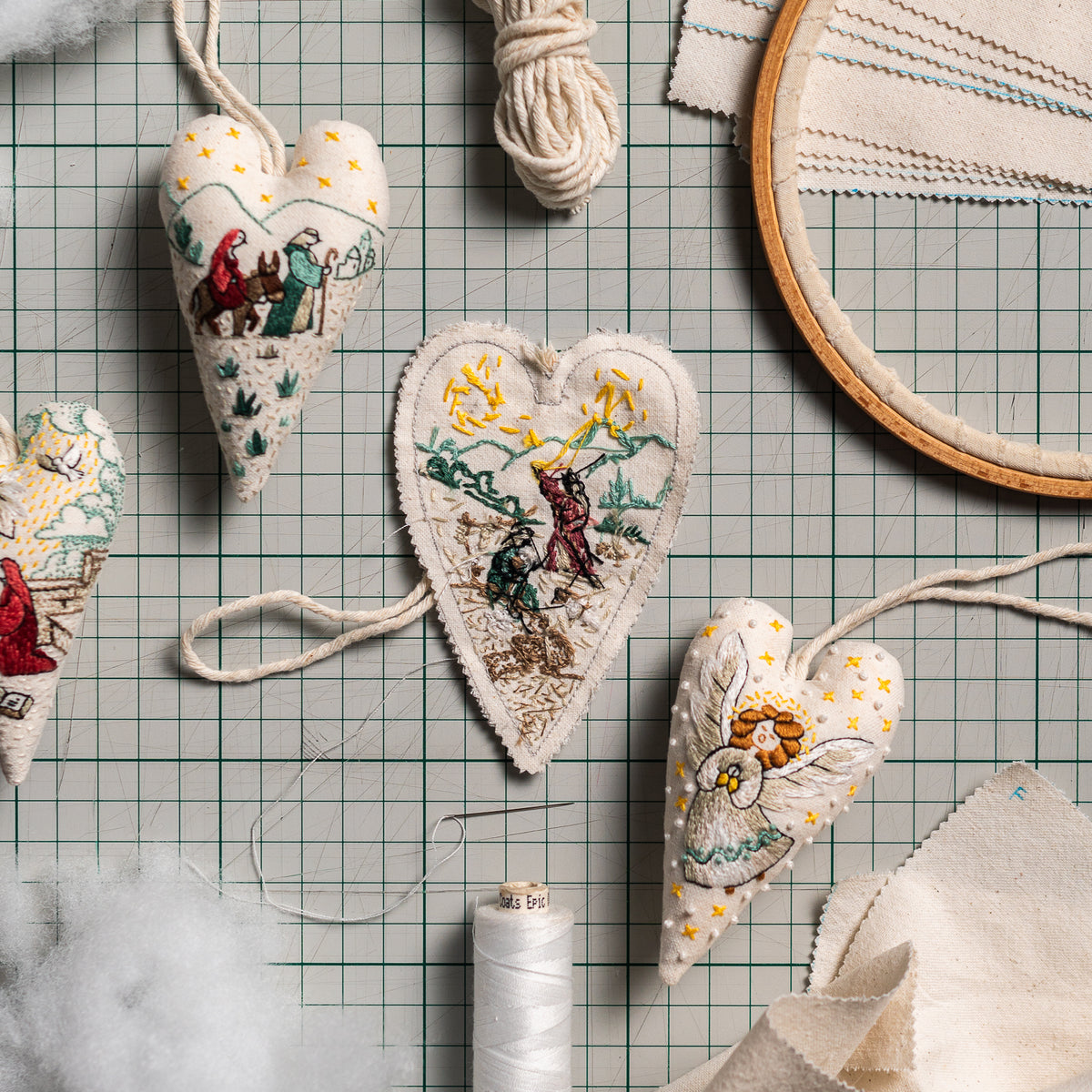 Handmade embroidered Christmas heart ornaments with Christian Nativity scenes arranged on a cutting mat alongside embroidery and sewing supplies.