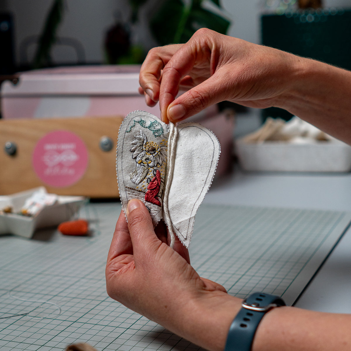 Hands assembling The Annunciation heart ornament, positioning the hanging cord between the embroidered front and fabric back.