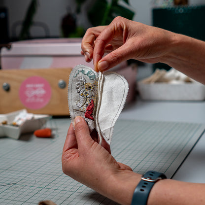 Hands assembling The Annunciation heart ornament, positioning the hanging cord between the embroidered front and fabric back.