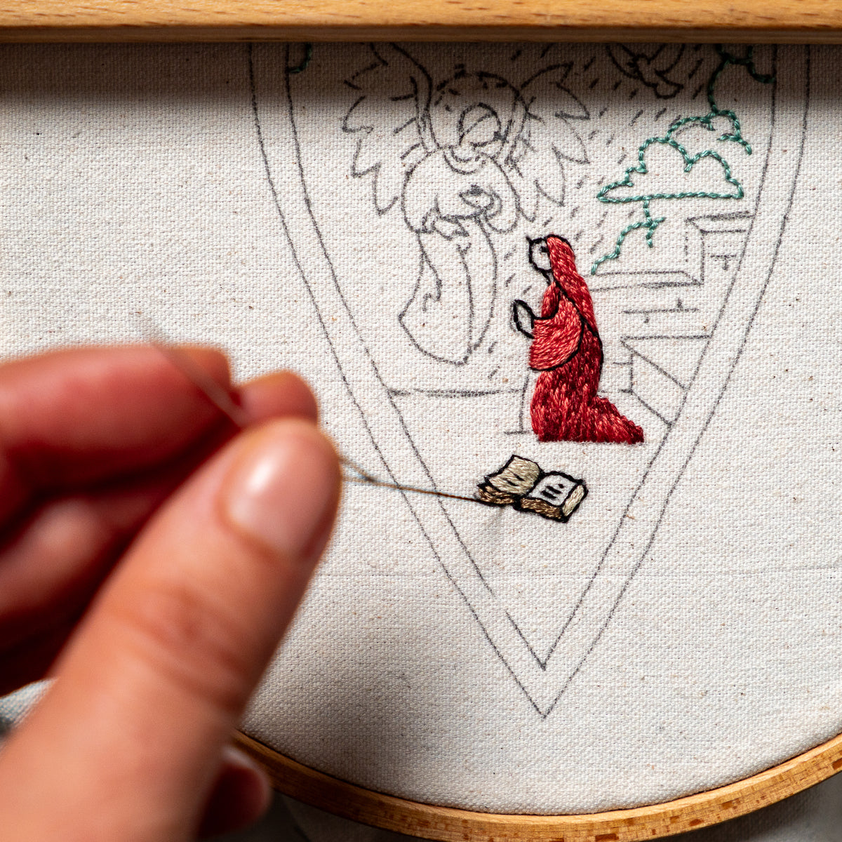 The Annunciation embroidery design in a wooden hoop, with Mary and a small book stitched onto linen fabric.