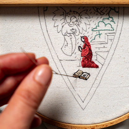 The Annunciation embroidery design in a wooden hoop, with Mary and a small book stitched onto linen fabric.