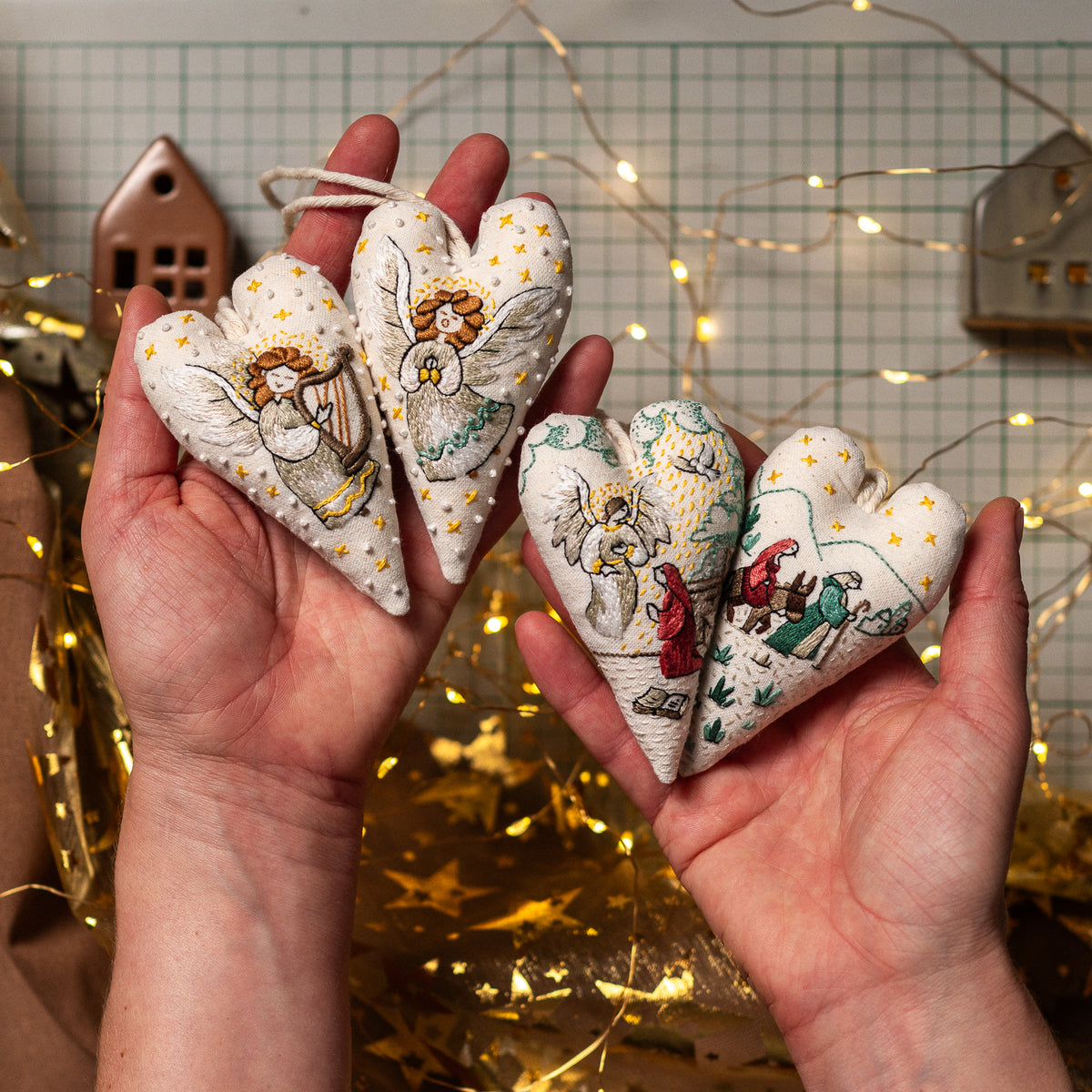 Hands holding several handmade Christmas heart ornaments featuring nativity embroidery designs, displayed against warm festive lights.