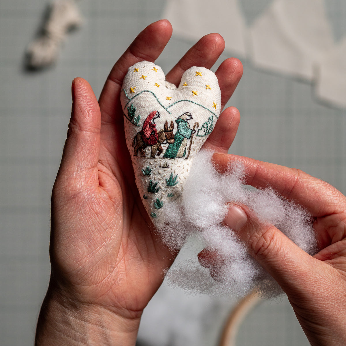 Hands adding soft polyester stuffing to a hand-embroidered nativity heart ornament featuring Mary, Joseph, and the donkey.