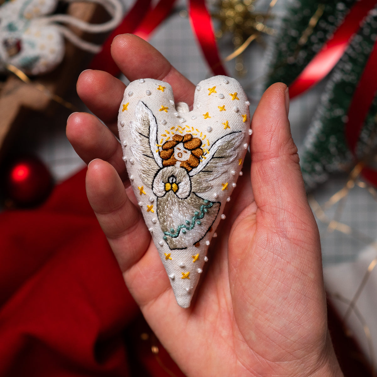 Close-up of a handmade herald angel heart ornament with silver embroidered wings, golden hair and tiny yellow stars, held in one hand.
