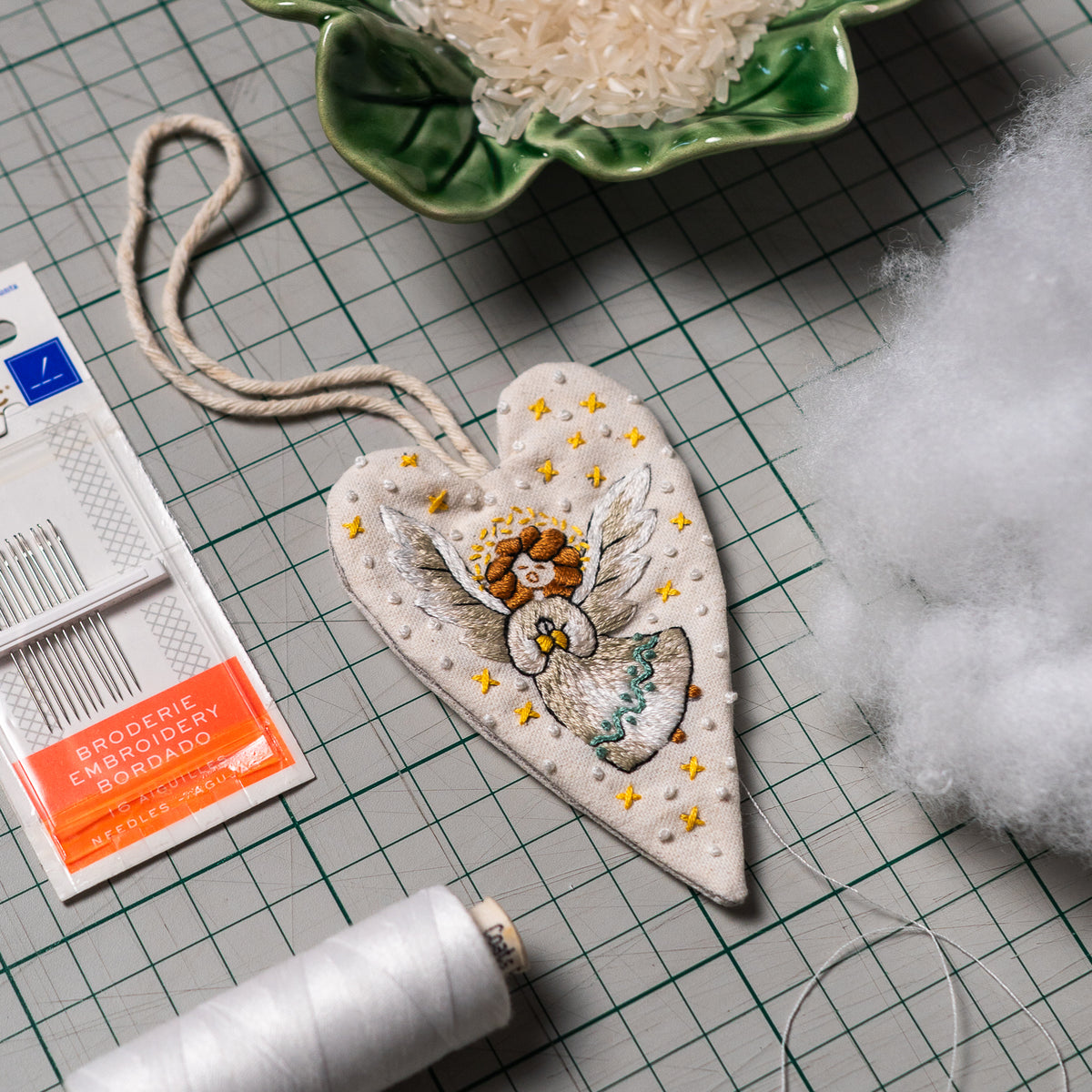 Hand embroidered angel heart ornament beside sewing needles, white thread, hanging cord and stuffing materials on a cutting mat.