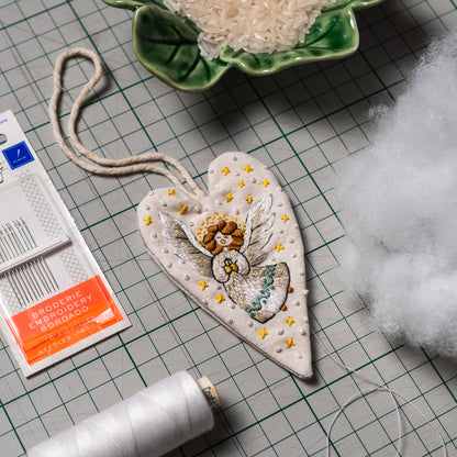 Hand embroidered angel heart ornament beside sewing needles, white thread, hanging cord and stuffing materials on a cutting mat.