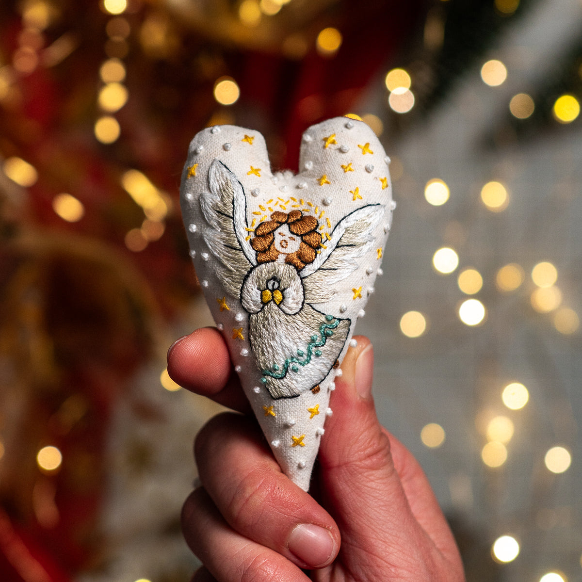 Hand embroidered Christmas heart ornament featuring a singing angel with golden hair, silver wings and yellow stars, held against warm Christmas lights.