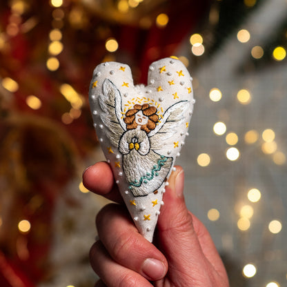 Hand embroidered Christmas heart ornament featuring a singing angel with golden hair, silver wings and yellow stars, held against warm Christmas lights.