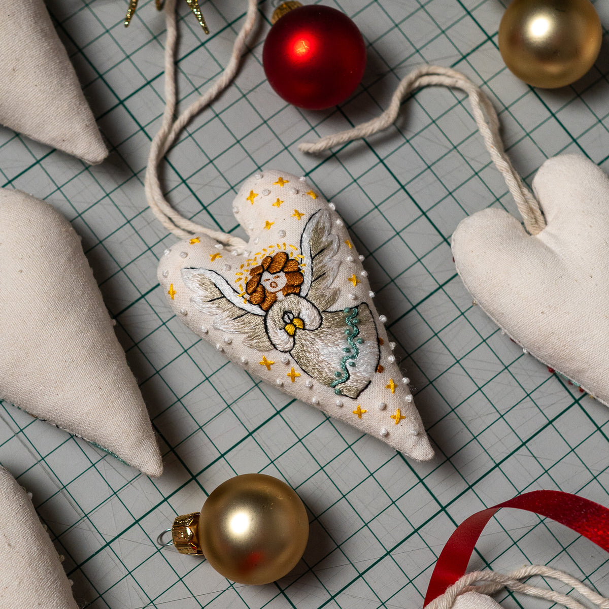 Finished angel embroidery heart ornament with a cream hanging cord, displayed among sewing materials and Christmas baubles.