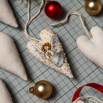 Finished angel embroidery heart ornament with a cream hanging cord, displayed among sewing materials and Christmas baubles.