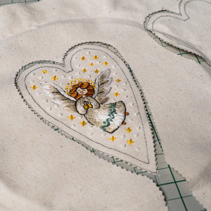 Embroidered herald angel design on heart-shaped linen fabric, cut with pinking shears and ready to sew into a Christmas ornament.
