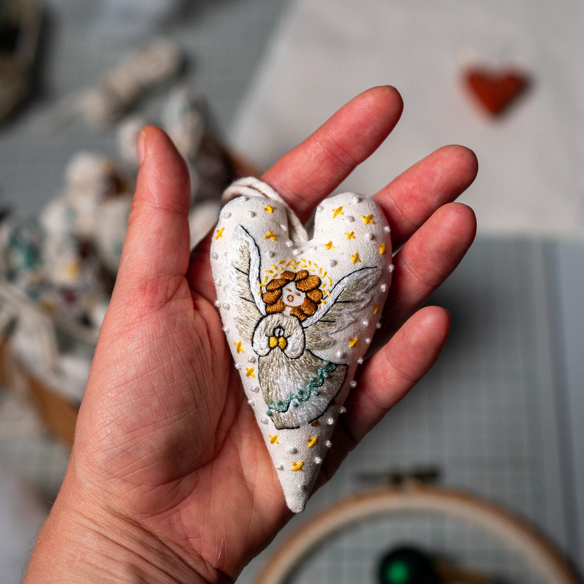 Finished hand embroidered herald angel Christmas heart ornament resting in an open hand, showing its silver wings and golden star details.