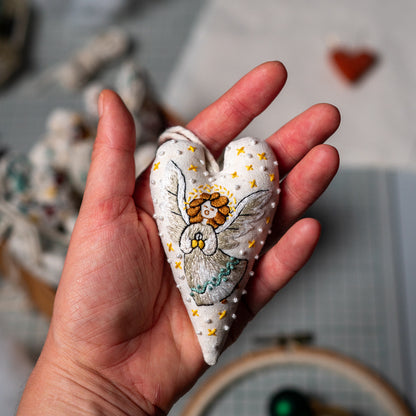 Finished hand embroidered herald angel Christmas heart ornament resting in an open hand, showing its silver wings and golden star details.