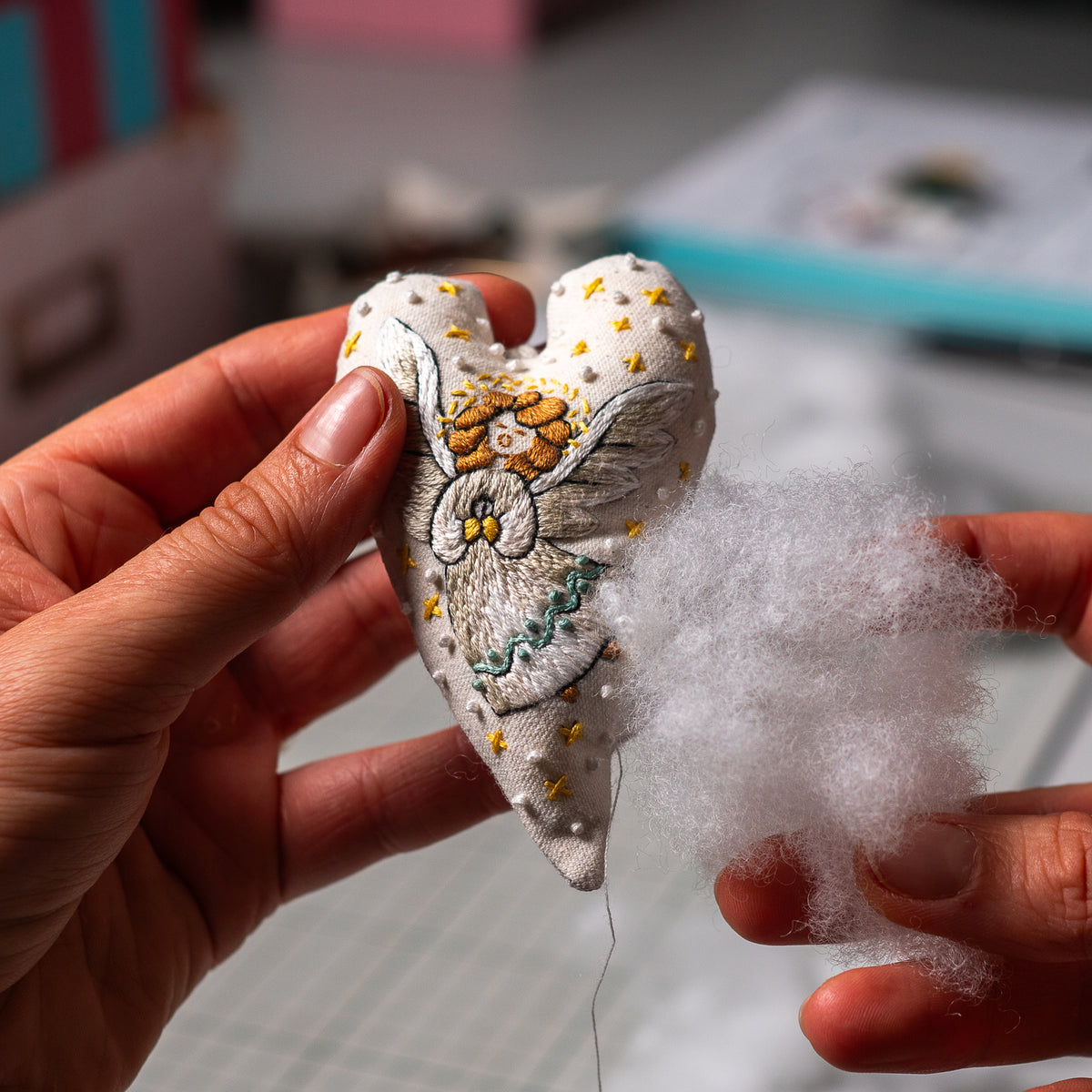Hands holding a partly sewn embroidered angel heart ornament and soft white stuffing during the Christmas ornament assembly process.