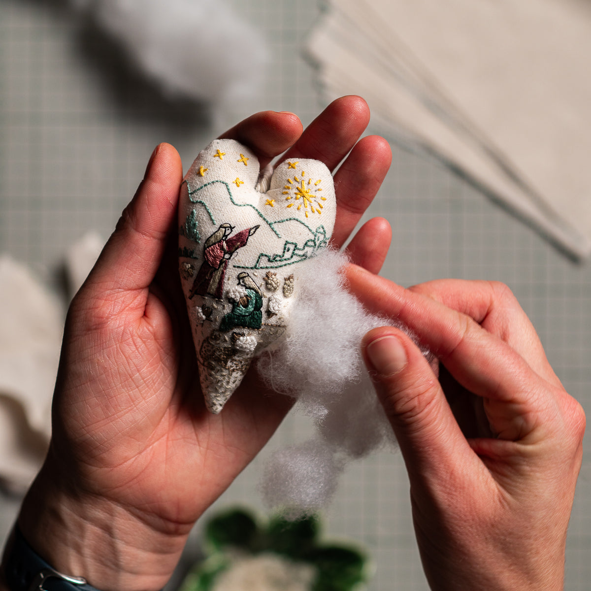 Hands adding polyester fiberfill to a Christian Christmas embroidered heart ornament featuring shepherds and sheep during the DIY sewing process.