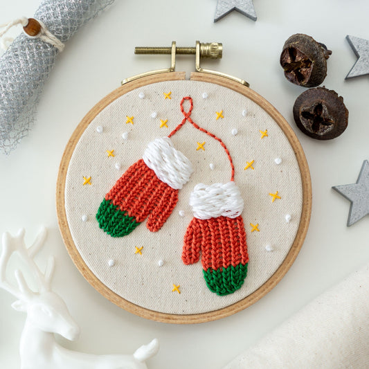 May include: Embroidered hoop art featuring two red and green mittens with white cuffs, surrounded by white dots and yellow stars on a beige background.
