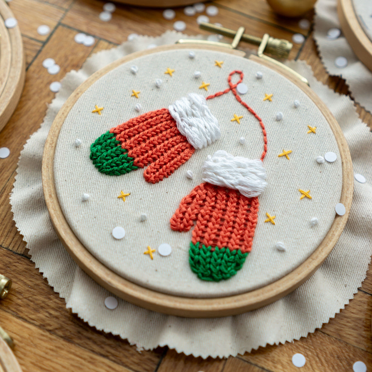 May include: Embroidered hoop art featuring a pair of red and green mittens with white cuffs, surrounded by white and gold stars on a white background.