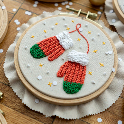 May include: Embroidered hoop art featuring a pair of red and green mittens with white cuffs, surrounded by white and gold stars on a white background.