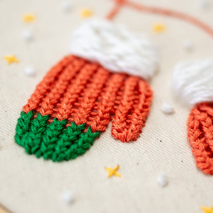 May include: Close-up of a hand-stitched embroidery design of two mittens. The mittens are orange and green with white cuffs. The background is a beige fabric with small white and yellow embroidered dots.