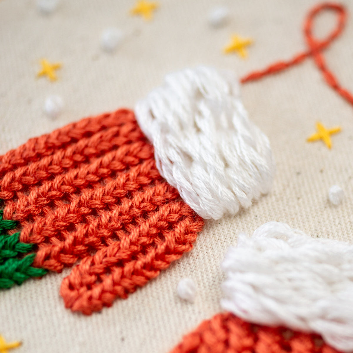 May include: Close-up of a hand-stitched embroidery design featuring a red and white knitted stocking with a green trim. The stocking is on a white fabric background with yellow and white embroidered stars.