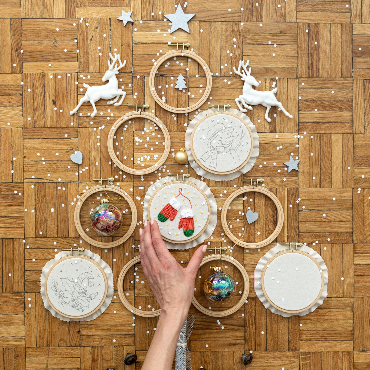 May include: A collection of 10 embroidery hoops with various Christmas-themed designs, including a snowman, mittens, a candy cane, and a Christmas tree. The hoops are arranged in a circle on a wooden surface.
