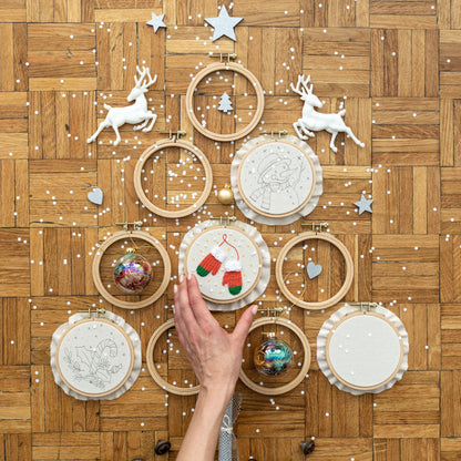 May include: A collection of 10 embroidery hoops with various Christmas-themed designs, including a snowman, mittens, a candy cane, and a Christmas tree. The hoops are arranged in a circle on a wooden surface.