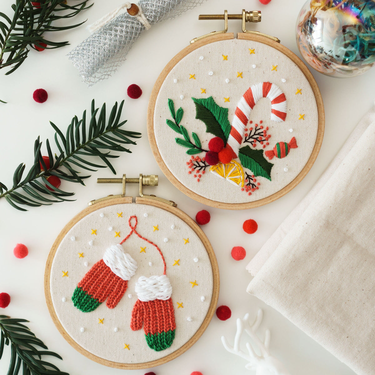 May include: Two embroidery hoops with white fabric and colorful embroidery designs. The top hoop features a red and white candy cane, green holly leaves, and a yellow lemon. The bottom hoop features two red and green mittens.