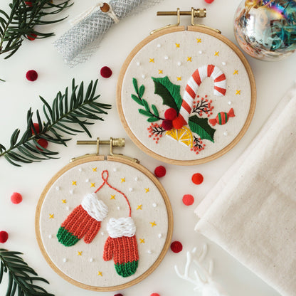 May include: Two embroidery hoops with white fabric and colorful embroidery designs. The top hoop features a red and white candy cane, green holly leaves, and a yellow lemon. The bottom hoop features two red and green mittens.