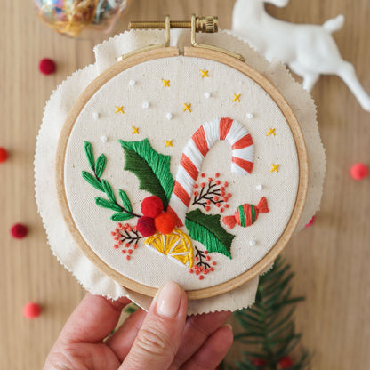 May include: A white embroidery hoop with a hand-stitched design of a candy cane, holly leaves, and a candy. The design is done in red, green, yellow, and white thread.