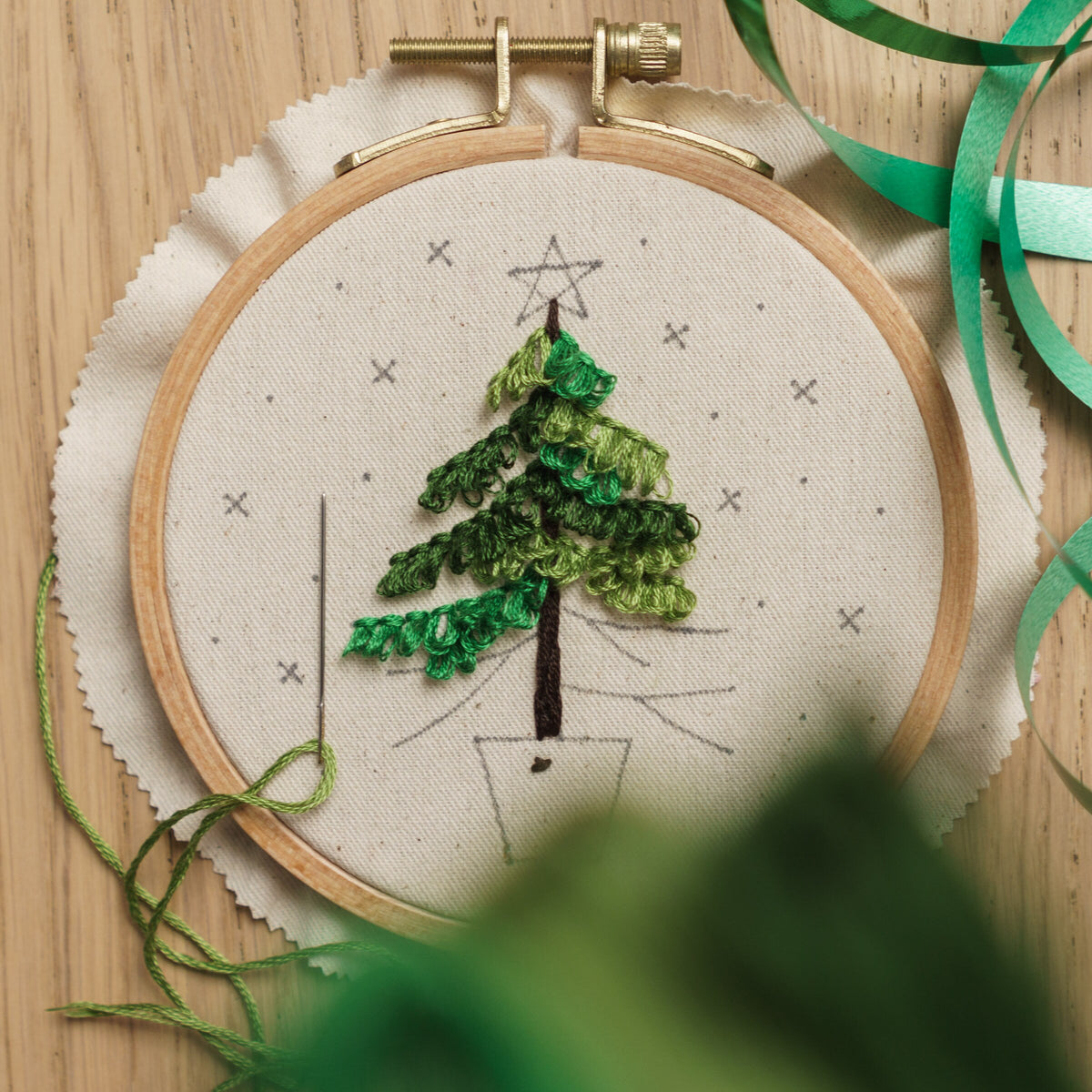 May include: A close-up of a hand-stitched embroidery hoop with a green Christmas tree design. The tree is surrounded by small white 'x' marks and a single star at the top. A green thread is threaded through a needle and is partially visible in the l