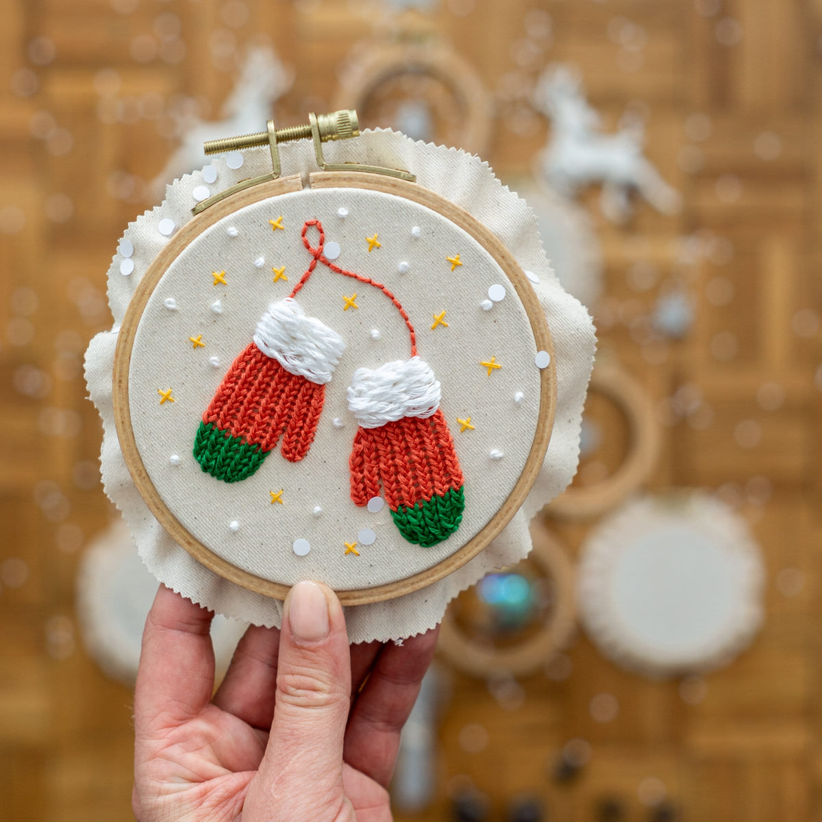 May include: A white embroidery hoop with red and green embroidered mittens on a white background with white and gold polka dots.