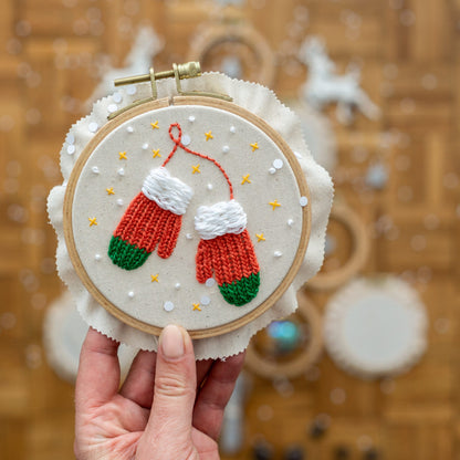 May include: A white embroidery hoop with red and green embroidered mittens on a white background with white and gold polka dots.