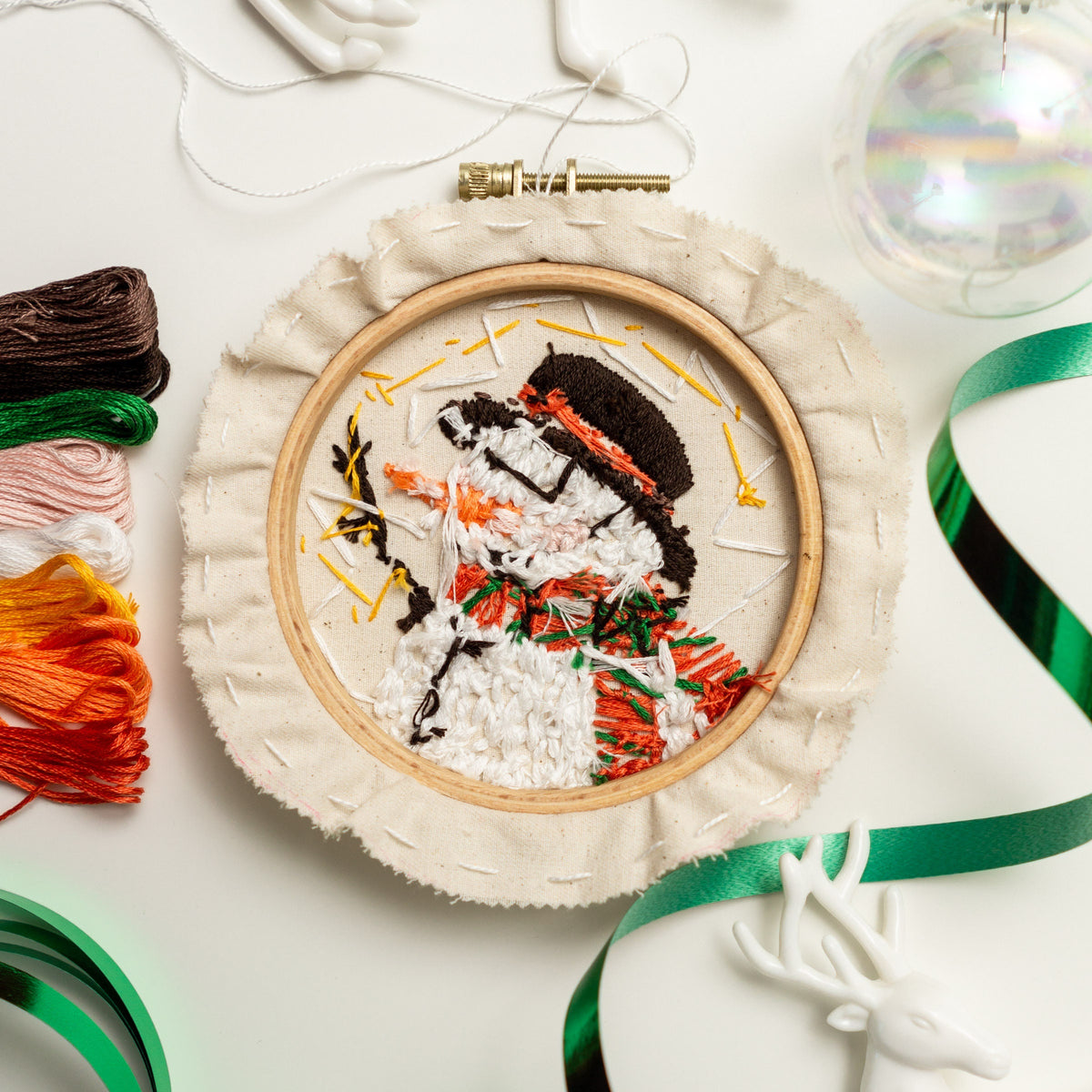 May include: A white embroidery hoop with a colorful embroidered snowman design. The snowman is wearing a red and green scarf and hat. The hoop is on a white surface with green and white ribbon.