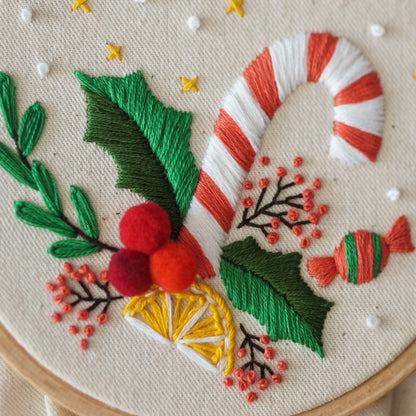 May include: Embroidered hoop art featuring a red and white candy cane, green holly leaves, red berries, and a green and red candy. The background is a natural linen fabric.
