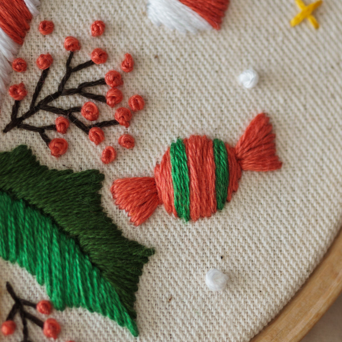 May include: A close-up of a hand-stitched embroidery design on a white fabric background. The design features a green holly leaf, red berries, and a red and green candy cane.