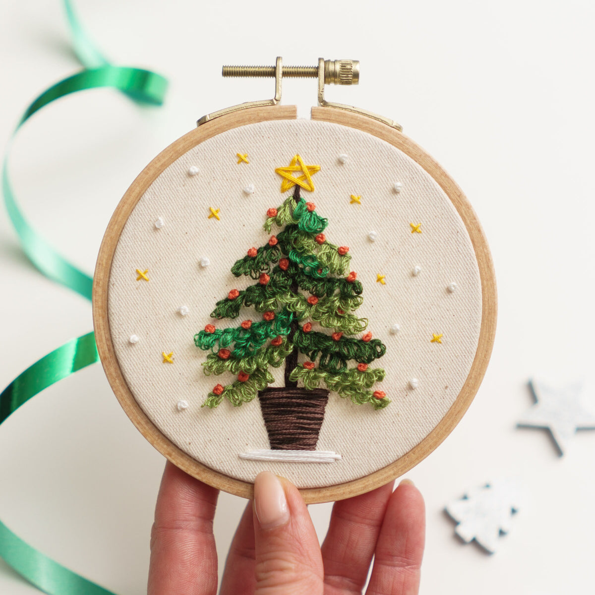 May include: Embroidered Christmas tree hoop art with a green tree, brown pot, and yellow star on a white background.